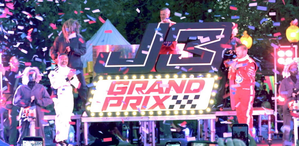 The on stage reveal of the J3 Grand Prix name logo. People in racing suits gather around a large logo and clap while confetti flies through the air.